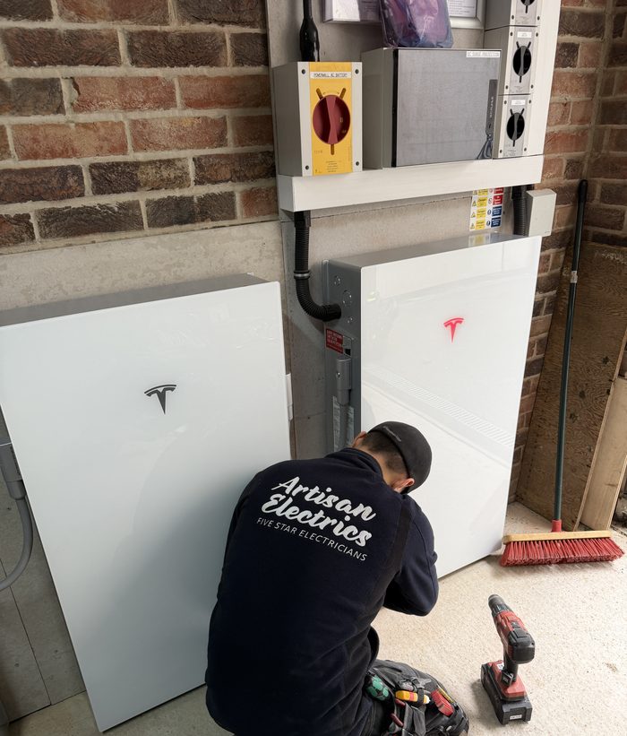 Artisan Electrics engineer kneeling in front of two Tesla Powerwall 3 units during DC Expansion installation — wearing branded Artisan Electrics Five Star Electricians uniform, power drill beside him, red Tesla logo visible on new Expansion Pack