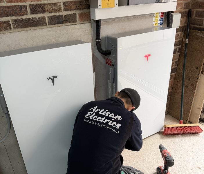 Artisan Electrics engineer in branded uniform kneeling beside Tesla Powerwall 3 and DC Expansion Pack during installation — power drill on floor, red Tesla logo on new Expansion Pack visible