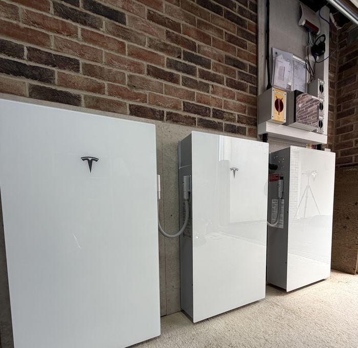 Three Tesla Powerwall 3 units side-by-side — one leader unit and two DC Expansion Packs — installed by Artisan Electrics in a UK home garage against an exposed brick wall, with electrical panel above